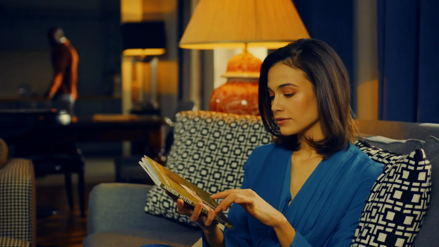 A woman reading a book on a sofa by lamplight in the evening